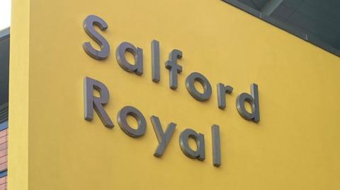 The Salford Royal Hospital sign. It is grey text emblazoned upon a yellow background.