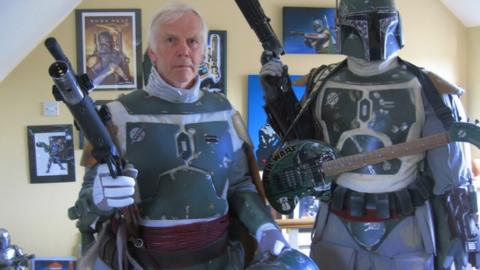 Jeremy Bulloch in Boba Fett costume standing alongside a Boba Fett figure, with Star Wars images on a wall in the background