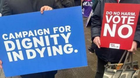 Two signs are held up, one is blue and reads "Campaign for Dignity in Dying" and the other is red and says "Do no harm. Vote no."