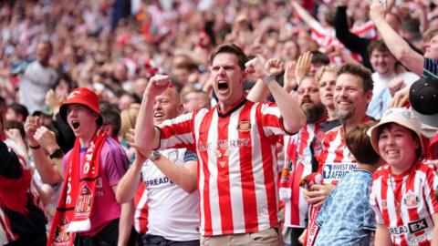 Group of Sunderland fans celebrating a goal. The man in the middle, wearing a Sunderland strip has both arms raised. Those around him -  also in Sunderland colours - are shouting or cheering.