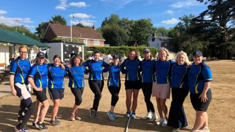 Eleven women in matching blue cricket uniforms stand together with their arms around one another. They're stood on a cricket pitch with a cricket house and scoreboard behind them.