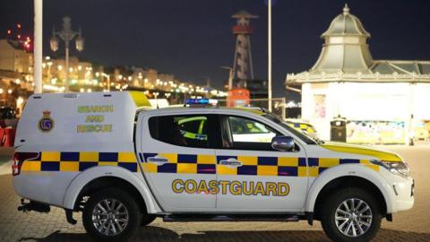A coastguard vehicle with blue and yellow markings is parked on Brighton seafront at night, with illuminated seaside buildings, streetlights, and a pavilion-style structure visible in the background.