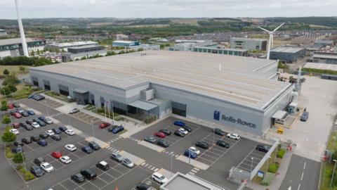 A large flat roof, single storey warehouse has the words Rolls-Royce in large blue letters along one wall. There is a car park outside