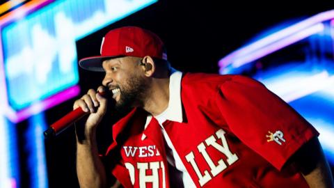 Will Smith sings into a red hand-held microphone. He is wearing a red cap and a red shirt which is open. 'West Philly' is written across the shirt in white. He is on a brightly-lit stage.
