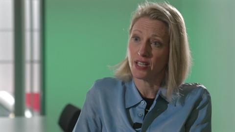 Claire Waxman, Victims' Commissioner, she has blonde shoulder length hair, and is wearing a blue shirt, sitting by a table and behind her is a green wall.