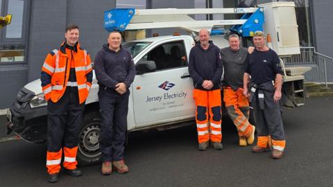 Jersey Electricity engineers are standing in front of a parked van. There are two men on the left, and three on the right. 