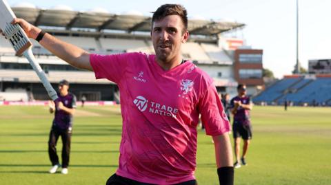 Craig Overton, wearing Somerset's pink shirt, celebrates the win over Yorkshire by raising his bat in the air.