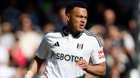 Rodrigo Muniz playing for Fulham. It looks like he is running and leaning to his left. His mouth is open. He is wearing a white Fulham home kit with the black Adidas logo on the left with the three stripes across the shoulders. The fans in the stand are out of focus behind him.