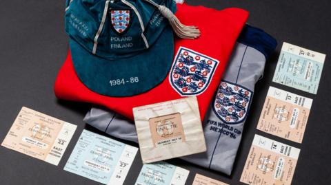 A pictures of a selection of tickets, Peter Shilton's cap with "Poland [and] Finland" sewed into the front of it and 1984-86 on the bill of the cap and two England badges, one sewed into a red shirt and another on a grey goalkeeper's top.