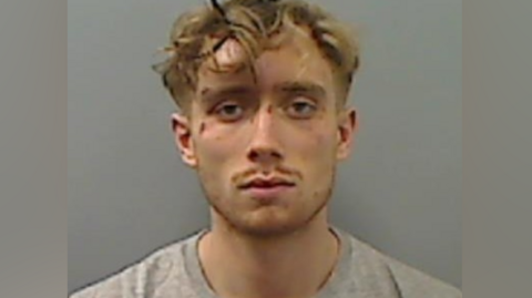 Mugshot of Callum Howe. He has blonde curly short hair and cuts to his face. He has a tiny moustache and is wearing a grey t-shirt.