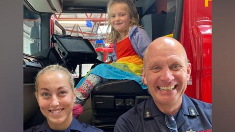 Olive sitting in the passenger seat of a red fire engine. She is smiling with two firefighters beside her.
