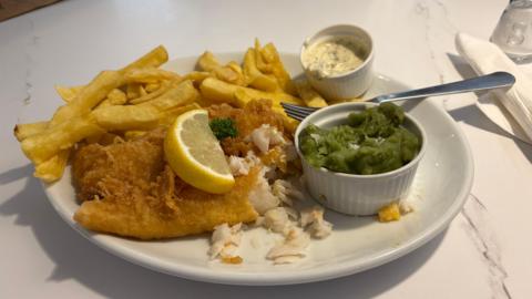 A plate of battered fish and chips, with a slice of lemon, a pot of mushy peas and tartare sauce. A fork is resting on the side of the plate. 