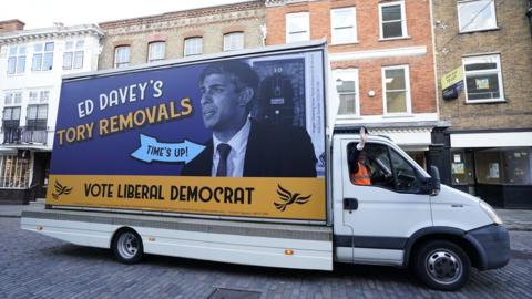 Sir Ed Davey driving a van through Guildford, unveiling a "Tory Removals" poster