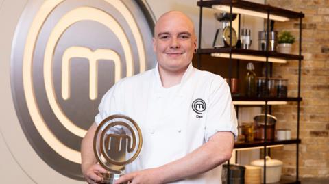 Dan Merriman in his white chef's jacket holds his silver Masterchef trophy in the studio with the circular Mastrchef logo and shelves of jars and pots behind him