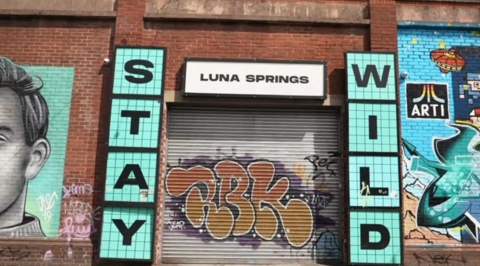 A brick wall painted with turquoise murals with the words Stay Wild in black lettering on a turquoise background on either side of a shuttered metal door covered in graffiti