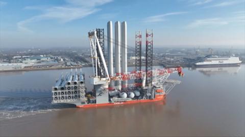 A massive offshore wind installation ship carrying turbine components sails along a river near an industrial port.