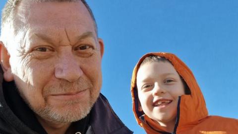 A father, in his late fifties, with his young son. Both are smiling into the camera. The child, who is around eight, is wearing an orange coat with his hood up.