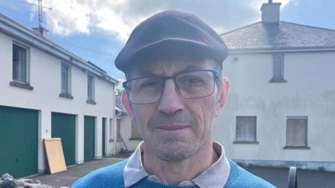 Sean Faloon standing in front of Clonduff parochial house on a sunny day in spring 2025.  He has short, greying dark hair and is wearing a dark flat cap, glasses and a blue jumper over a polo shirt.  