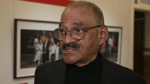 Ramon "Sugar" Deen is standing in an exhibition in front of framed photographs. He has very short hair and black moustache. He is wearing dark framed glasses.