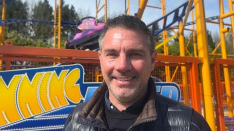 A man standing in front of a colourful amusement‑park ride, with bright yellow and blue coaster tracks and a pink‑and‑purple car behind him, smiling while wearing a dark jacket on a sunny day with trees in the background.