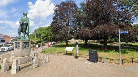 A town centre park with large trees with a pavement around the outside, and a small black railing separating the pavement and the park. There is an information board alongside the railing. A statue of the author John Bunyan coated in green patina can be seen to the left of the picture on a stone plinth, with small concrete posts at the base. There is a road to the left of the statue.