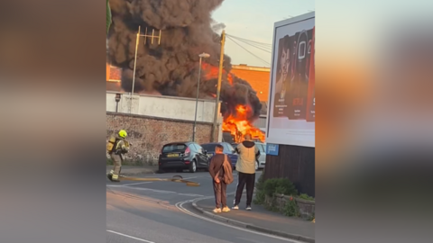 Two men gaze at a fire in the distance as a firefighter walks towards it. 
