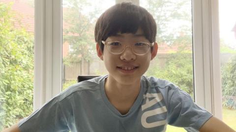 A boy with short black hair, glasses and a blue shirt, smiles as he sits in a conservatory. A back garden can be seen through the window behind him.