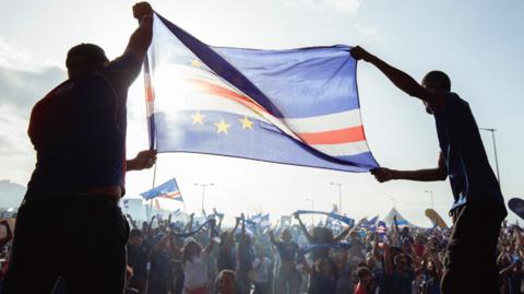 Two men silhouetted hold aloft a Cape Verde flag which is rendered almost translucent as sunlight shines through it as hundreds of people celebrating are seen in front of them waving more flags and holding up scarves 
