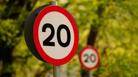 Two 20mph signs with trees in the background. One of the signs is blurred. The signs are on two poles.