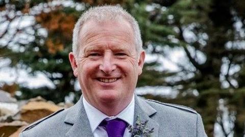 Roland Crichton is a middle aged man with short grey hair. He is standing outside and there are trees in the background. He is wearing a grey Highland dress jacket over a white shirt and a purple tie.