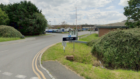 A road junction into Long Lartin Prison with a black and white Long Lartin sign in the grass near a bush. A car park can be seen in the background along with multiple brick buildings.