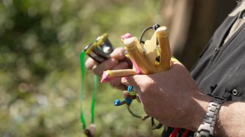 Close up picture of hands holding catapults in a woodland setting.  