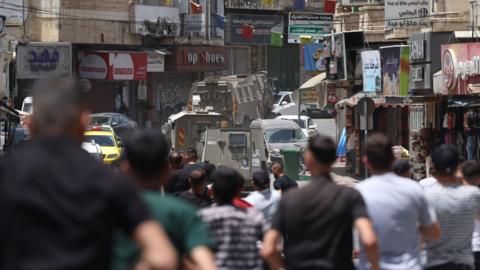 Palestinian men are standing on a street in Nablus on 27 May. Israeli tanks are in the background while shops are at the side