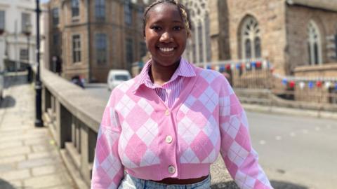 A woman in a pink patterned top with a number of buildings behind her