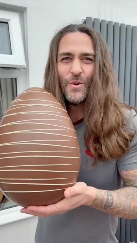 Billy D'Souza looks at the camera. He has long brown wavy hair and wears a grey short sleeved t shirt. He is holding a giant milk chocolate Easter egg with white chocolate stripes on it.