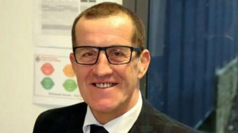 A man with short cropped dark hair and wearing a suit and glasses smiles at the camera