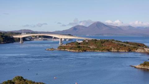 Skye Bridge pictured on a bright, sunny day. Behind the bridge is a range of hills, and the crossing spans calm, blue waters.