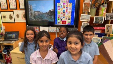 Shows five students (three girls and two boys) smiling at the camera stood infront of their artwork. Their piece is alongside Canaletto's Regatta on the Grand Canal, which inspired the piece. Their classroom walls are filled with art.