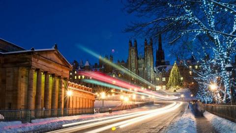 Light trails from traffic moving along a snowy road in central Edinburgh, with an illuminated Christmas tree visible in the distance.