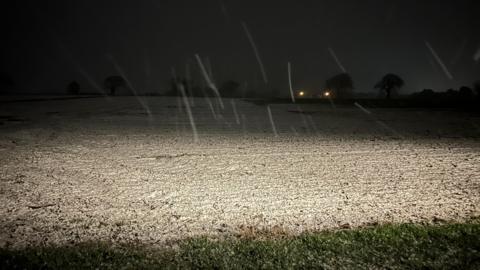 A night-time image of a lit up field with a light scattering of snow. Rising up in the distance are a few trees on the horizons and two lights, possibly from houses.