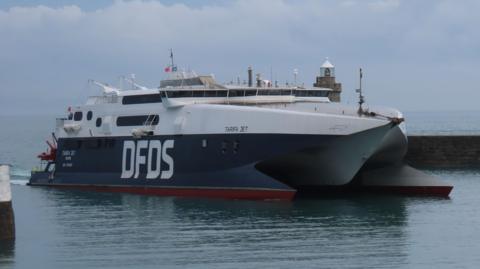 DFDS ferry Tarifa Jet travels into a harbour on a cloudy day. The vessel is half dark blue and half white. It has the DFDS logo on its side written in large white block capitals on the dark blue section of the boat.