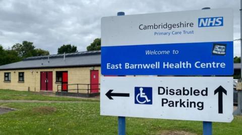 NHS sign which says "East Barnwell Health Centre" with a sign underneath saying disabled parking. A single storey building with beige bricks and red doors is behind it. 