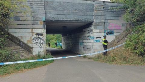 A police cordon is put in place in front of a bridge with a police officer stood looking away 