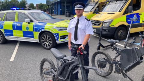 Insp James May, in his police uniform of a black cap, white shirt and black tie, stands behind a black electric bike. There are police patrol cars and yellow police vans in the background. 