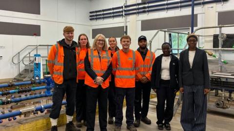 A group of young men and women pose for a photo with Baroness Jacqui Smith - wearing hi-vis jackets and standing in front of blue pipes and scaffolding