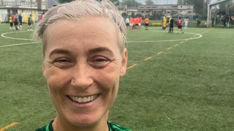 Women looking directly at the camera with light, short blonde hair with a green football field behind her.