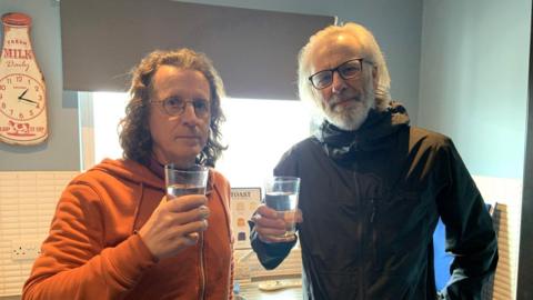 A man with brown shoulder length hair is wearing glasses and an orange hoodie. He is standing next to a man with shoulder length grey hair and a grey beard wearing a black jacket and glasses. Both men are in a kitchen holding glasses of water