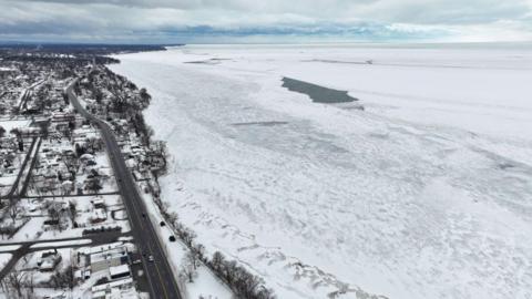 Aerial view of the shoreline of Lake Erie. Roads, houses and snow-covered land on one side, a white covered lake on the other.