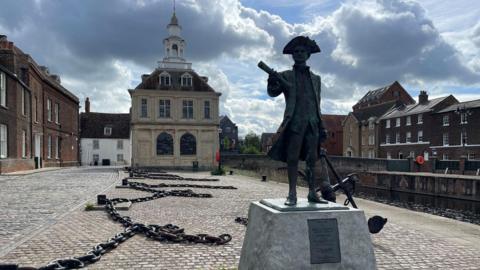 Ship chains lay along a quayside. Buildings are across the water and a statue of a man wearing a tricorn three-cornered hat stands on a grey, stone plinth.