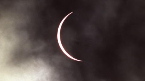 The moon has moved across the sun leaving a thin band of the sun visible which has taken on a pink hue. The sky is cloudy.